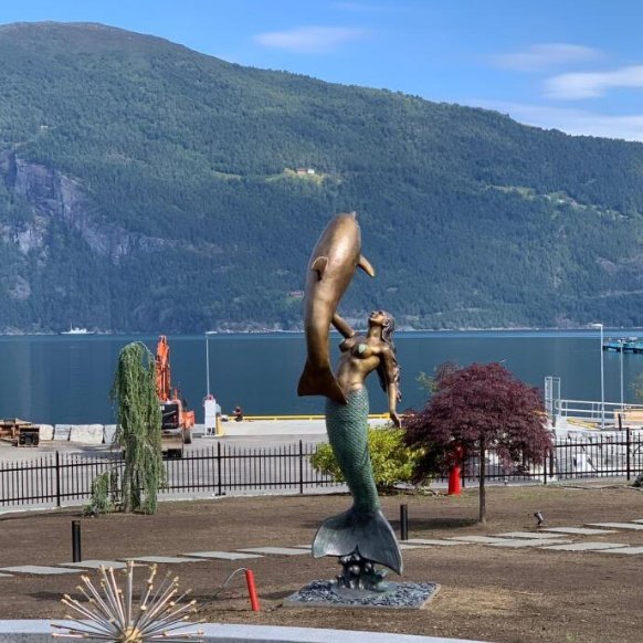 Monumental bronze mermaid and dolphin sculpture in a coastal landscape in Norway